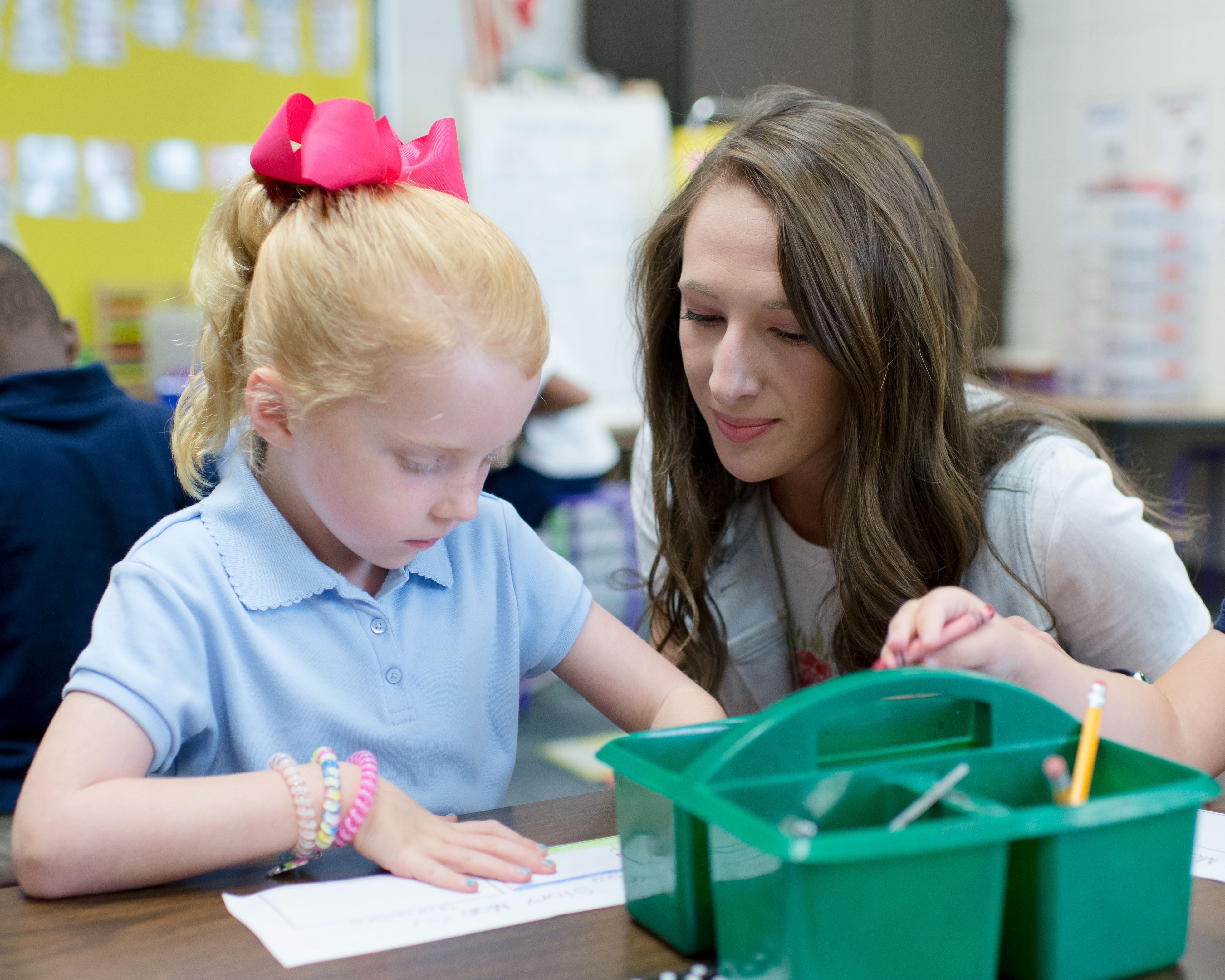 Teacher observing a student, who wears a pink bow in her hair, at her desk.