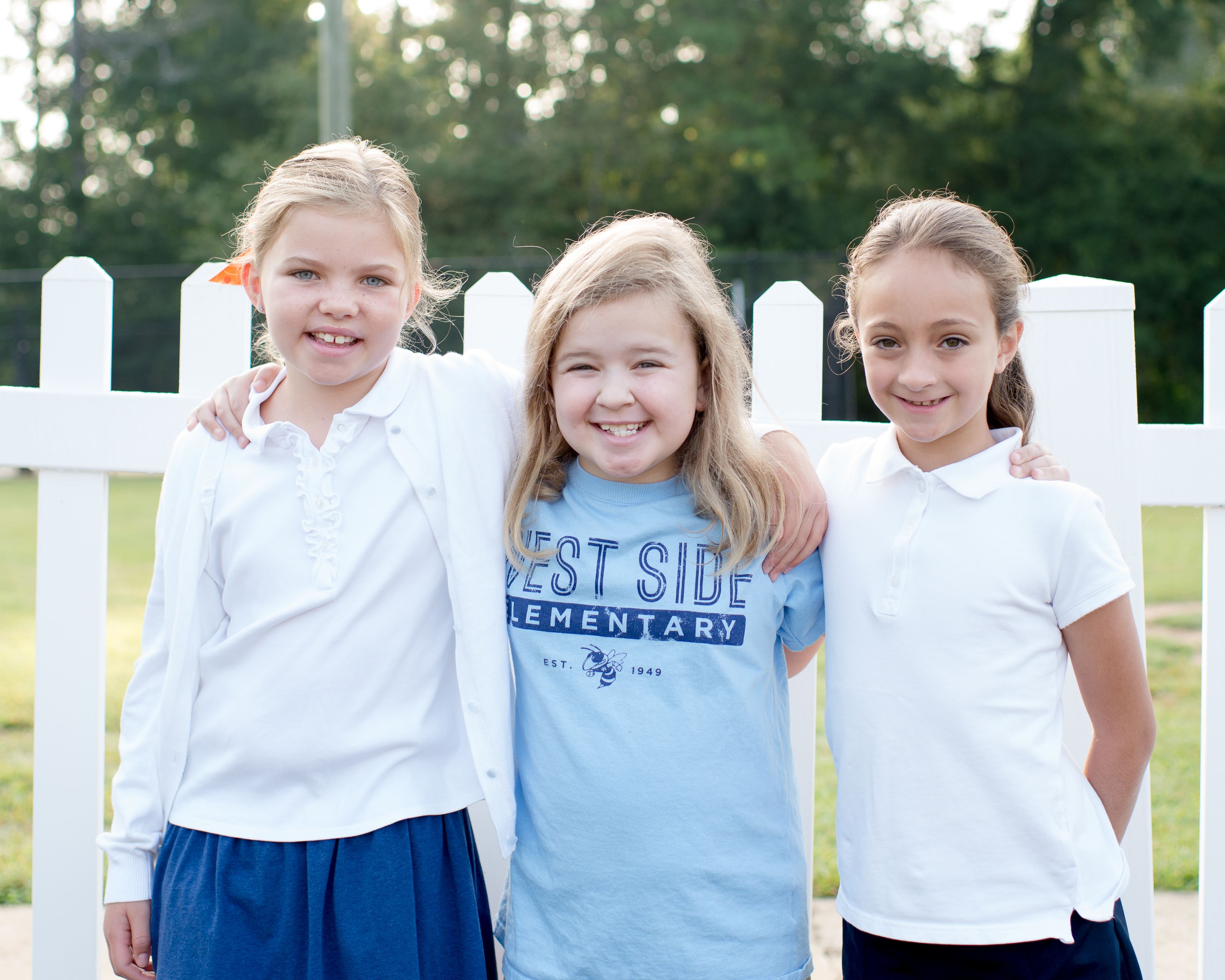 Three girls smiling together in front of white picket fence, the center student in a blue West Side t-shirt Three girls smiling together in front of white picket fence, the center student in a blue West Side t-shirt