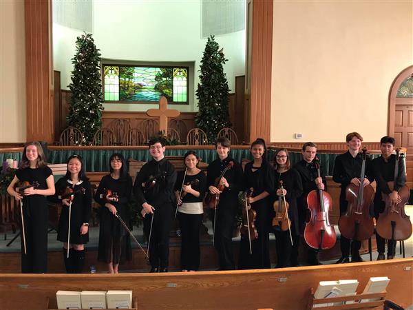 Orchestra students holding instruments and smiling in a church with cross and Christmas trees behind them