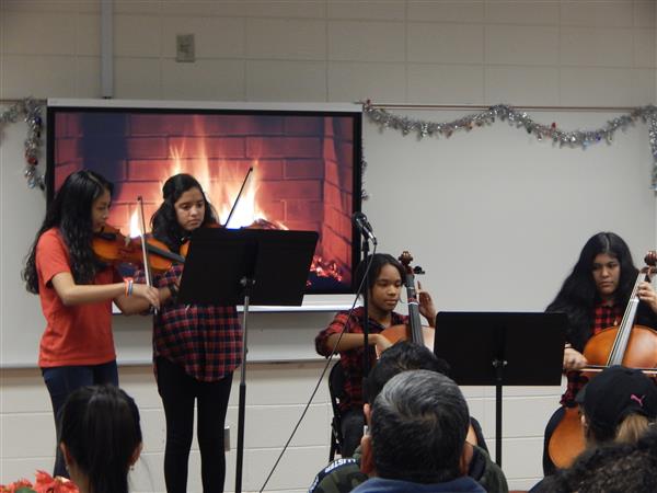 Four students play string instruments for classroom full of adults