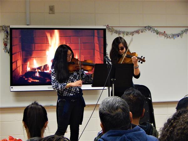 Two students play string instruments in front of classroom full of adults