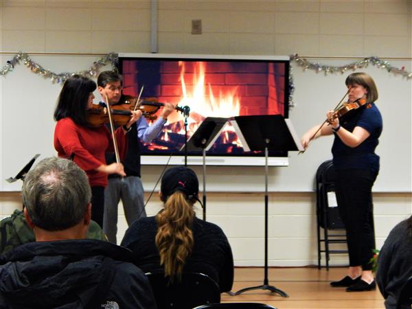 Three adults play string instruments in front of classroom of adults