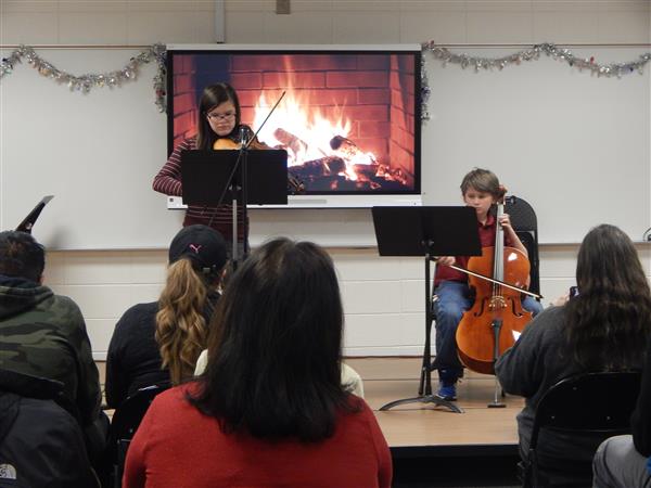 Two students play string instruments in classroom with adults in audience