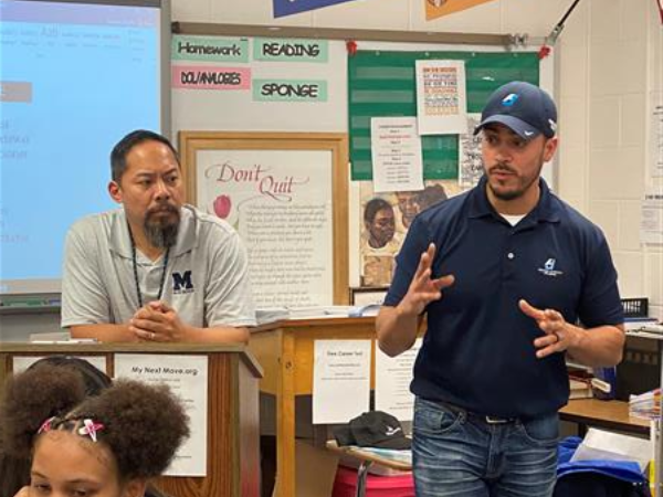 Man in grey shirt beside man in blue hat and jacket addressing students in classroom