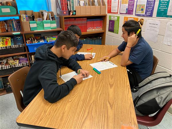 Three students with highlighters and papers at table in classroom