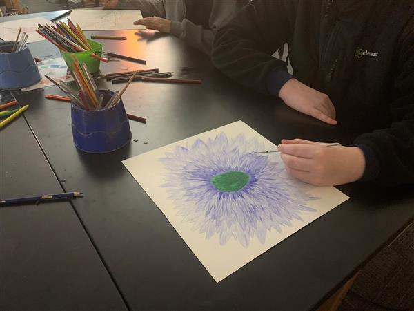 Close-up of student painting a blue flower with green center in watercolors
