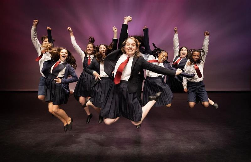 A group of students in costume for Matilda jump on stage with their fists in the air