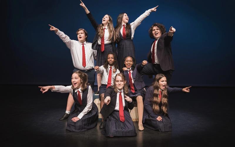 A group of students in costume for Matilda stand at varying levels on stage and point in different directions