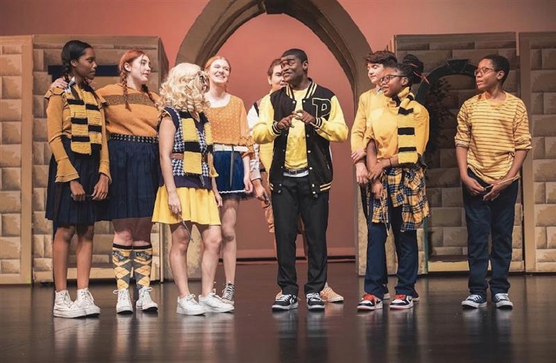 Students dressed in yellow costumes stand on the stage in the middle of their performance for Puffs