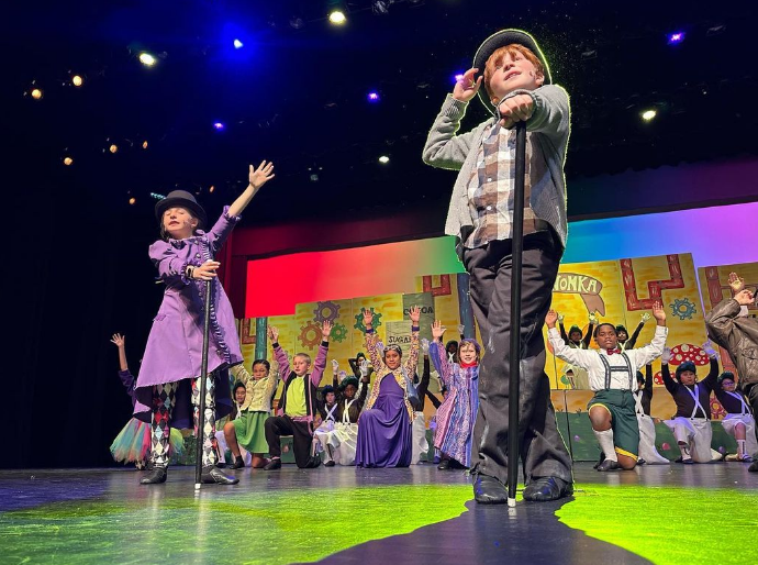 Two young students in costume for Willy Wonka Jr stand close to the camera with the rest of the cast behind them