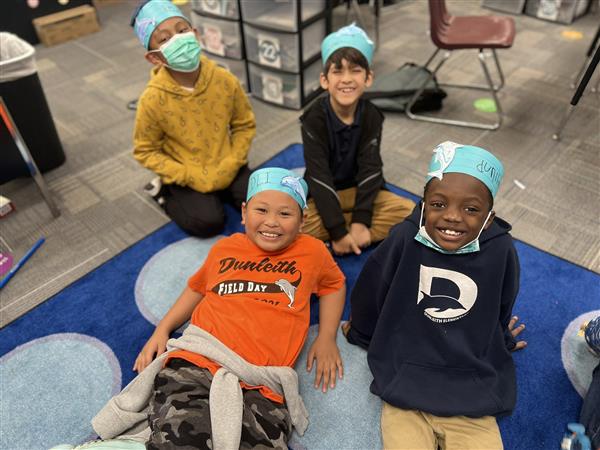 Students in paper Dunleith hats sit on the floor and smile