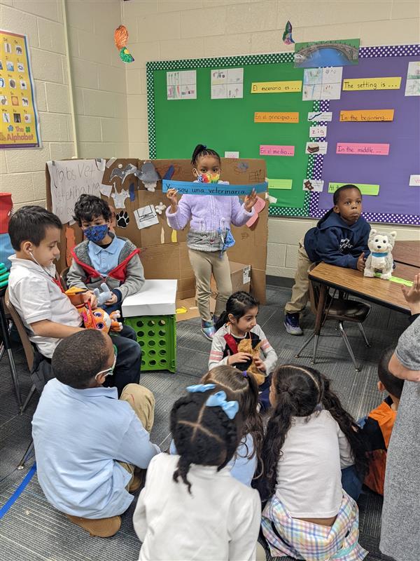 Students sit on floor while another presents a project in Spanish