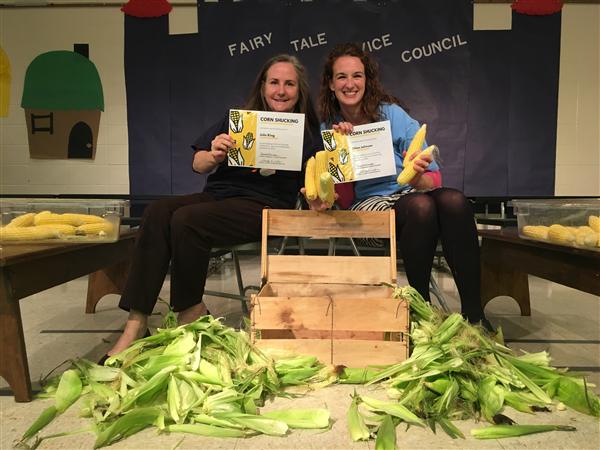 Two women in front of empty crate, surrounded by shucked corn cobs and husks, holding certificates from a shucking competition