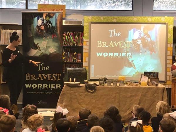 Women addressing seated children with display presenting book called the Bravest Worrier