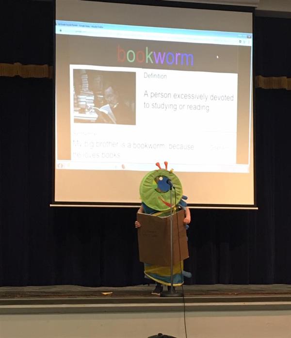 Child in bookworm costume standing on stage, with slide behind defining bookworm as a person excessively devoted to studying or reading