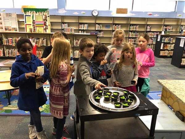 Students excitedly grabbing small cups of a green snack from a round silver tray in library