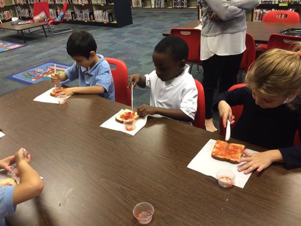 Students seated at table practice spreading portions of red jelly onto slices of bread with plastic knives