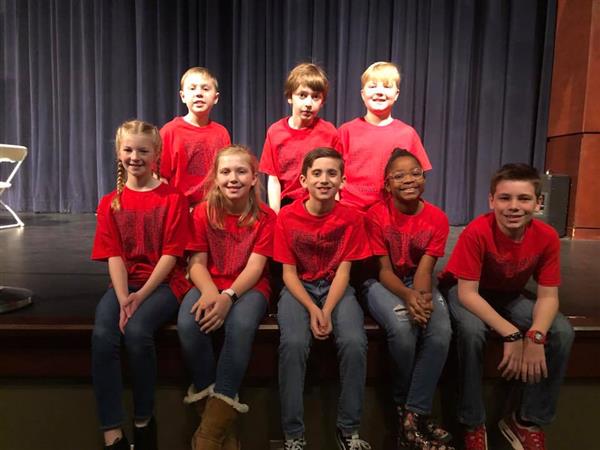 Eight students pose together, wearing matching red shirts, sitting on edge of stage in auditorium