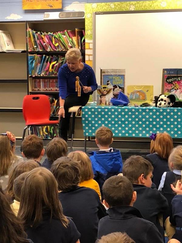 Woman with horse marionette addresses seated students, with books displayed on table beside her